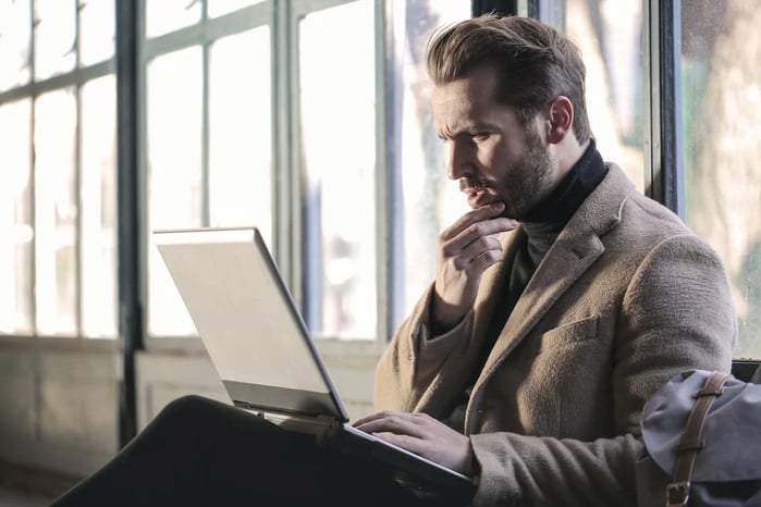 A man in a coat looking at a laptop on his lap with a confused expression.